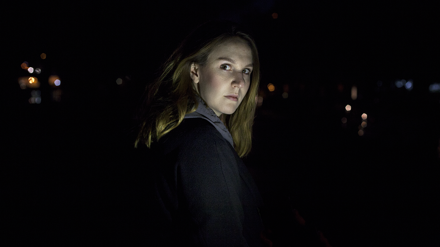 Surrounded by darkness, woman looks over her shoulder, down the barrel of the camera with concern