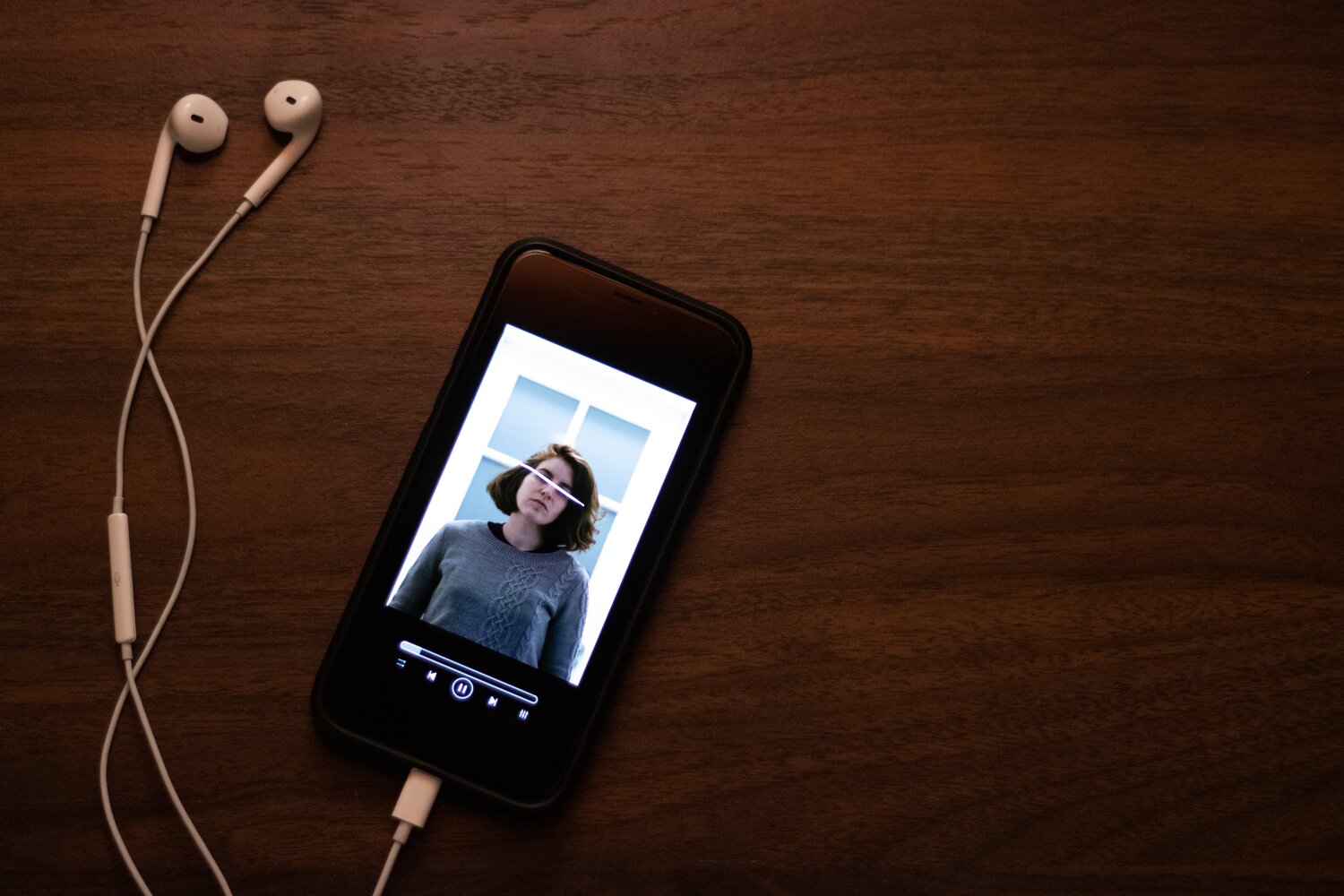 A phone and headphones sit on a wooden surface. The display depicts an audio track is playing and the wallpaper shows a photo of a woman will her eyes censored.