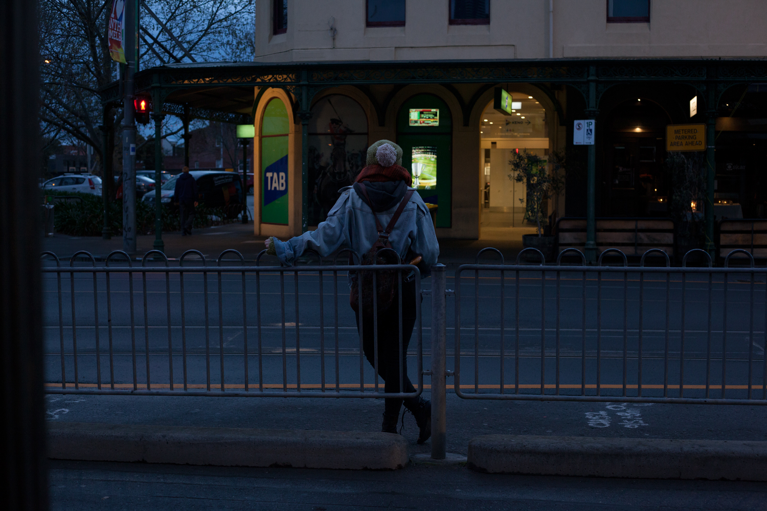 At dusk, a woman stands waiting at a tram stop with her back to us