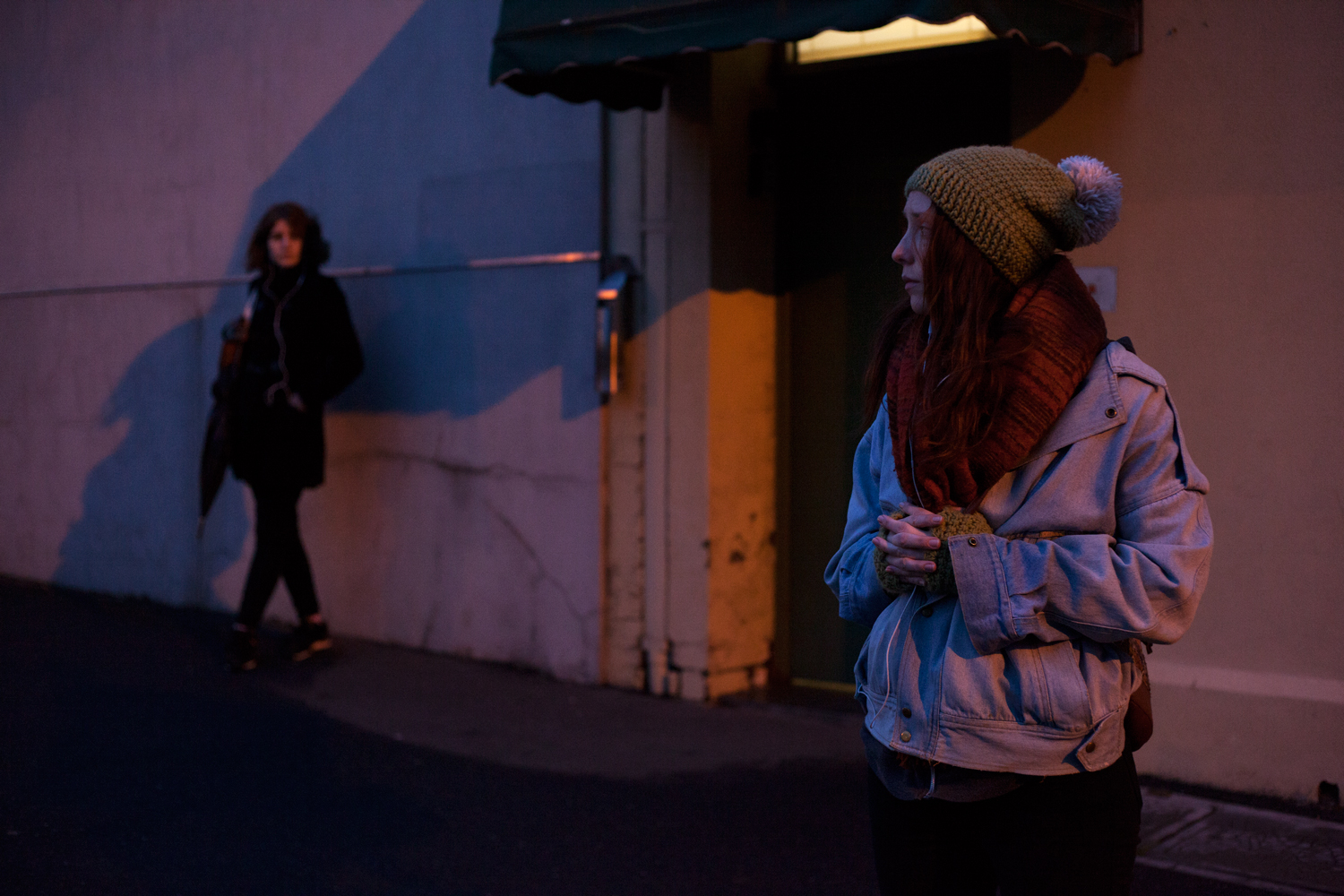 A woman stands on the street at dusk, and looks nervously out of frame. In the background, out of focus, another woman watches