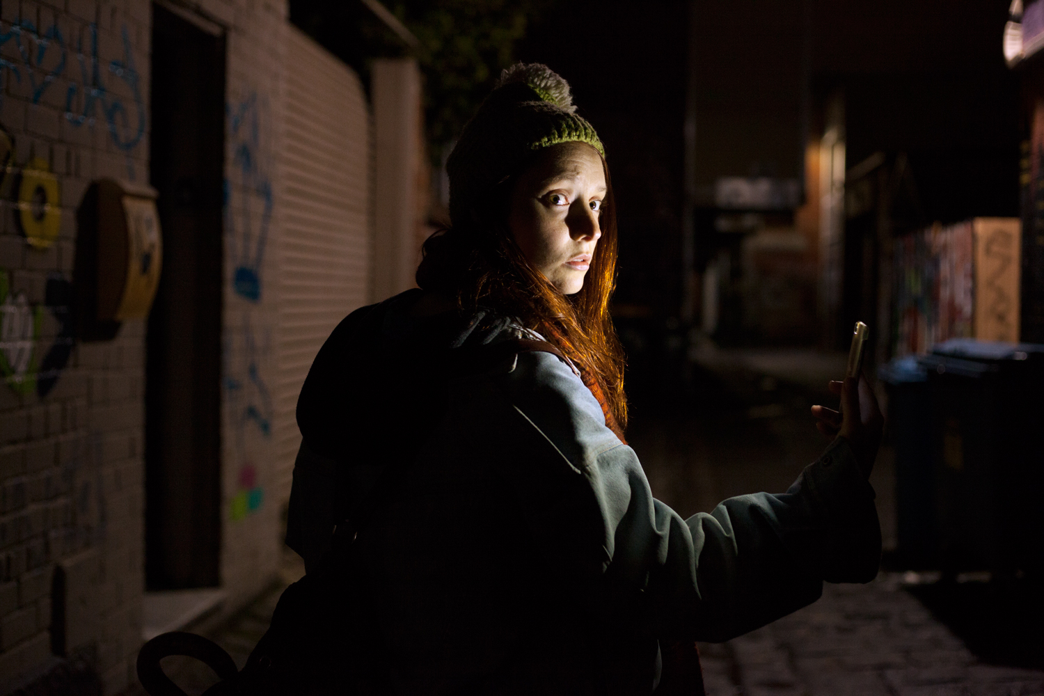 A woman in a dark alley looks over her shoulder, worried; her phone illuminates her face.